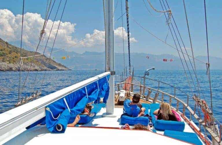 A group of people enjoying a sunny day on the deck of a sailboat with a clear view of the sea and mountains in the distance.