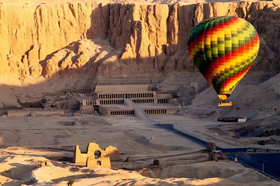 Colorful hot air balloon floats over the Temple of Hatshepsut in Luxor, Egypt, with sunlit cliffs in the background.