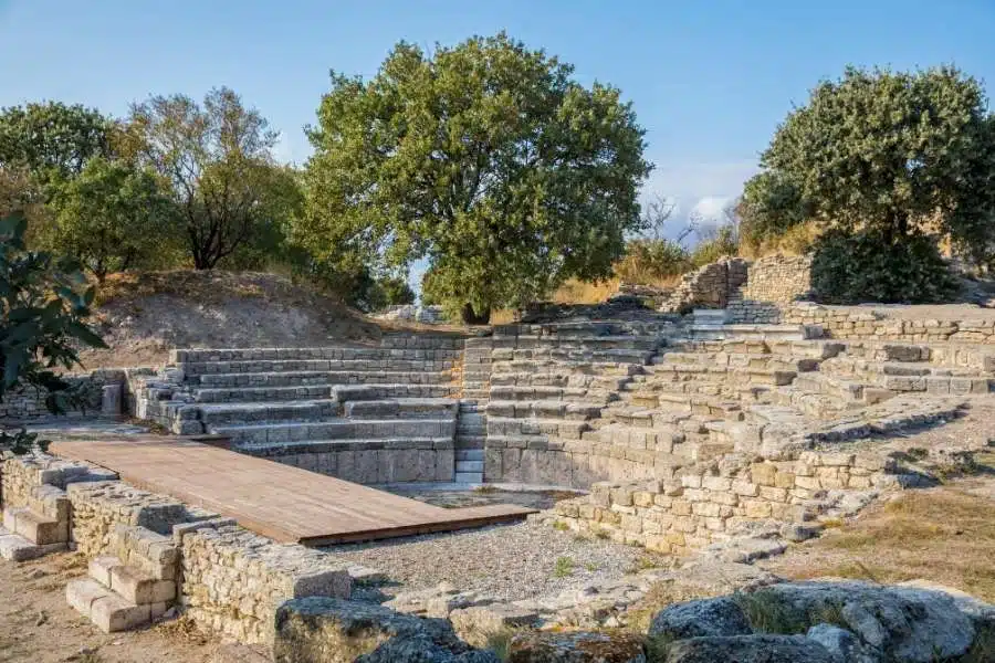 Stone seating of the Odeion and Bouleuterion in ancient Troy, surrounded by trees and sunlight, with a wooden platform.