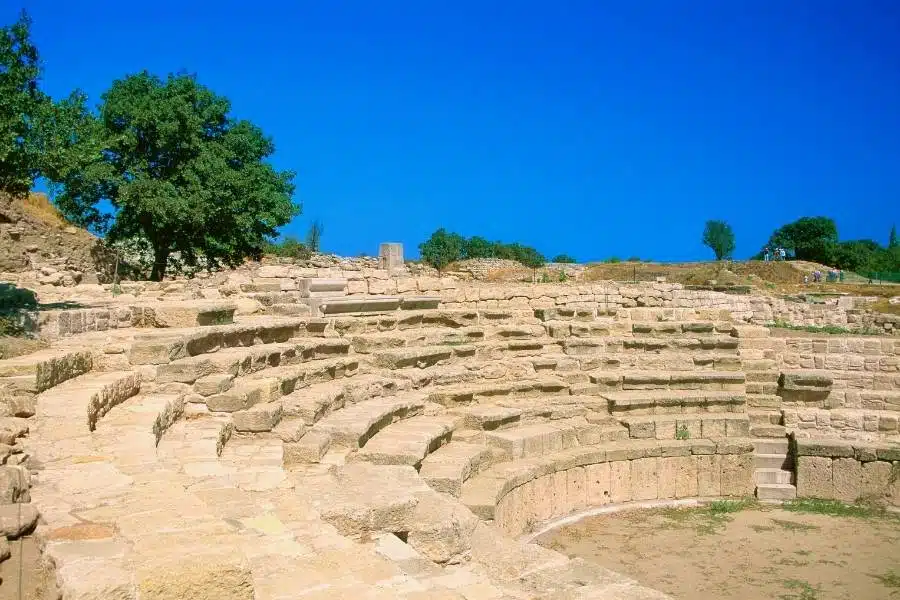 Stone seating of the ancient Odeon amphitheater ruins in Troy, Turkey, under a bright blue sky with trees in the background.