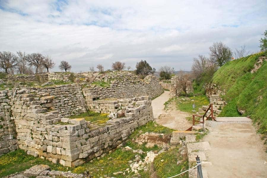 Stone ruins in the ancient city of Troy, Turkey, with grassy paths, low walls, and scattered greenery under a cloudy sky.