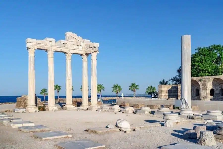 Ruins of the Temple of Apollo in Side, Turkey, with tall columns and a coastal view framed by palm trees and blue sky.