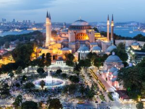 Illuminated Hagia Sophia in Istanbul at dusk, with vibrant city lights, minarets, and a lively park in the foreground.