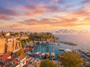 A scenic sunset view of the historic Old Harbor in Antalya, Turkey, featuring ancient stone walls, red-roofed buildings, a marina filled with wooden boats, turquoise waters, and distant snow-capped mountains under a warm, colorful sky.