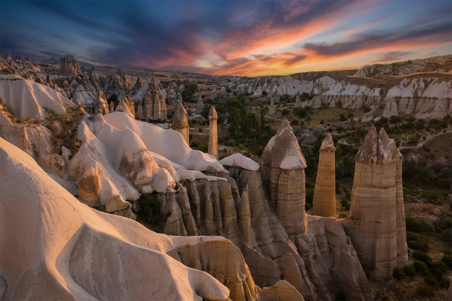 A breathtaking sunset view of the iconic fairy chimneys and ancient volcanic rock formations in Cappadocia, Turkey, illuminated under a vibrant orange and purple sky.
