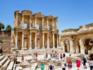 A group of ten tourists walking in front of the historic Library of Celsus in Ephesus, Turkey, under a clear blue sky.
