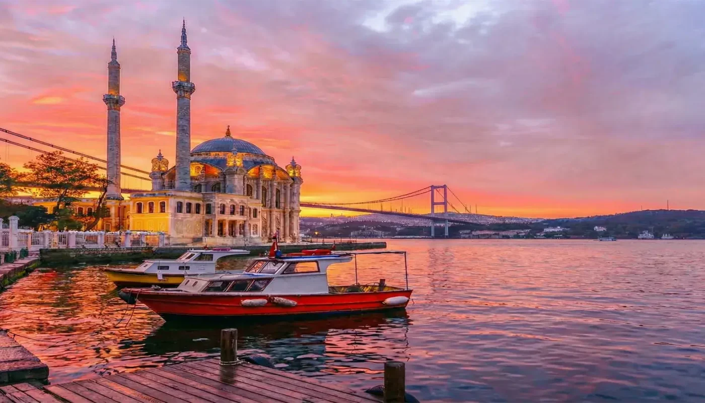 Experience the magic of Istanbul at dusk. This breathtaking photograph captures the illuminated Ortaköy Mosque and the Bosphorus Bridge spanning the strait against a dramatic, colorful sunset sky, with traditional boats in the foreground.